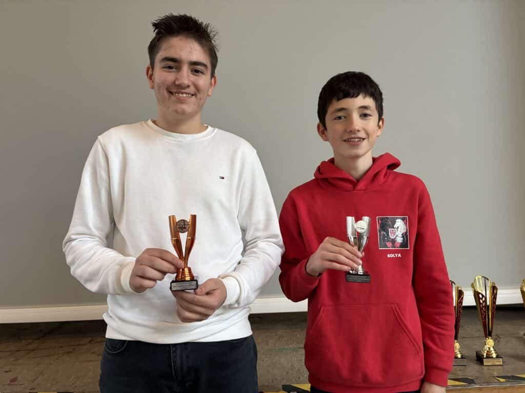 Zwei lächelnde Jungen stehen Seite an Seite und halten kleine Trophäen in der Hand, während größere Trophäen auf einem Tisch hinter ihnen vor einer schlichten hellen Wand stehen.