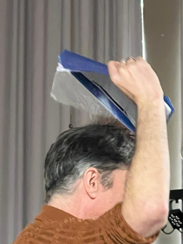 Eine Person in einem braunen Hemd hält eine blaue Mappe über den Kopf, als wolle sie sich damit schützen oder einen Schlag auf den Kopf vorbereiten.