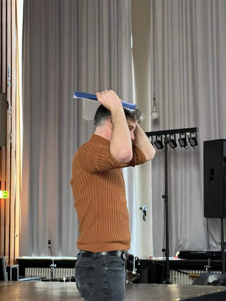 Ein Mann in einem braunen Pullover steht seitlich und hält einen blau-weißen Gegenstand über seinen Kopf in einem Raum mit hohen, grauen Vorhängen und Bühnenausstattung im Hintergrund.