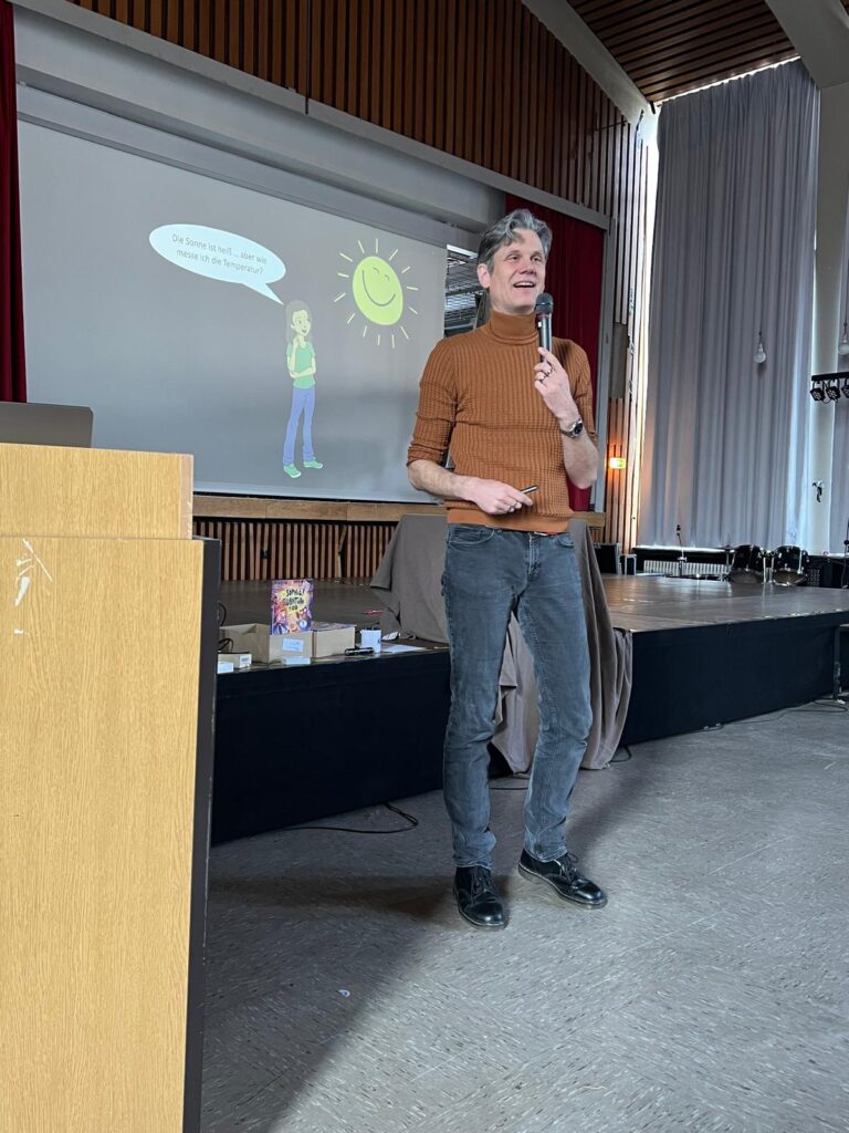 Eine Person in einem braunen Pullover hält ein Mikrofon in der Hand und spricht auf der Bühne vor einem projizierten Cartoon-Bild einer lächelnden Sonne und eines Menschen in einem großen Raum.