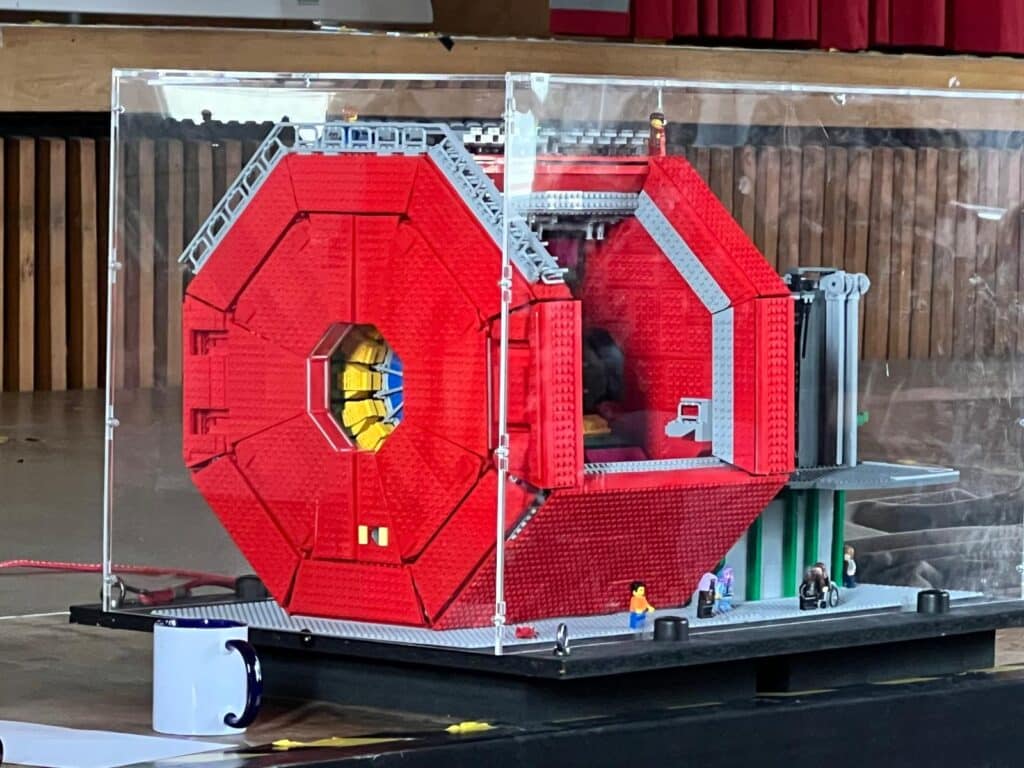 Ein detailliertes LEGO Modell einer großen, roten, sechseckigen wissenschaftlichen Maschine ist in einem durchsichtigen Gehäuse ausgestellt, in dessen Nähe kleine LEGO Menschenfiguren stehen, um den Maßstab zu wahren.