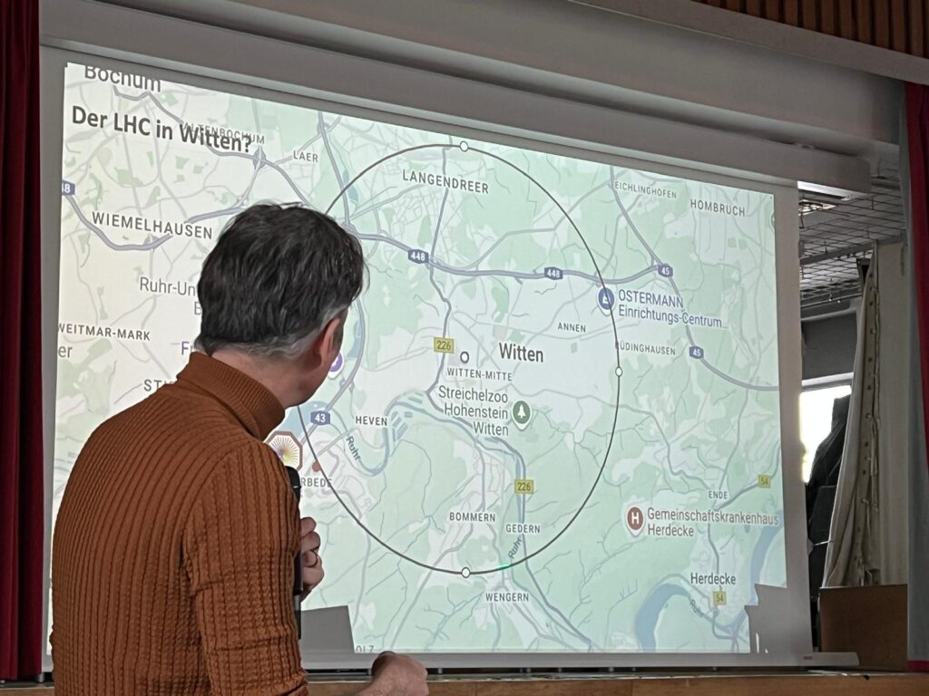 Ein Mann in einem braunen Pullover spricht in ein Mikrofon, während er auf eine projizierte Karte zeigt, die ein kreisförmiges Gebiet um Witten, Deutschland, mit der Aufschrift Der LHC in Witten? zeigt.