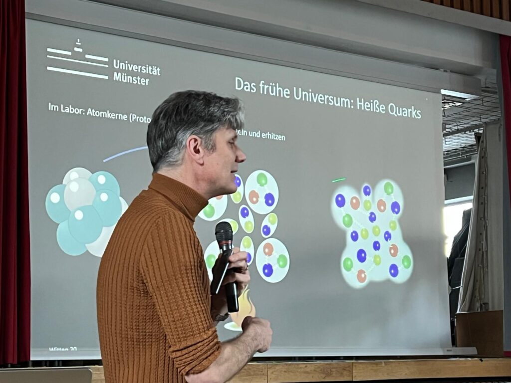 Ein Mann in einem braunen Pullover hält ein Mikrofon in der Hand und präsentiert eine Folie über Das frühe Universum: Heiße Quarks mit bunten Atomdiagrammen an der Universität Münster.
