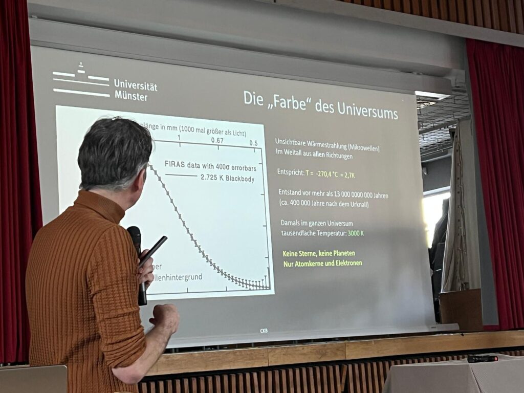 Eine Person präsentiert an der Universität Münster in einem Klassenzimmer mit roten Vorhängen eine Folie über die Farbe des Universums mit einer Grafik und einem deutschen Text.