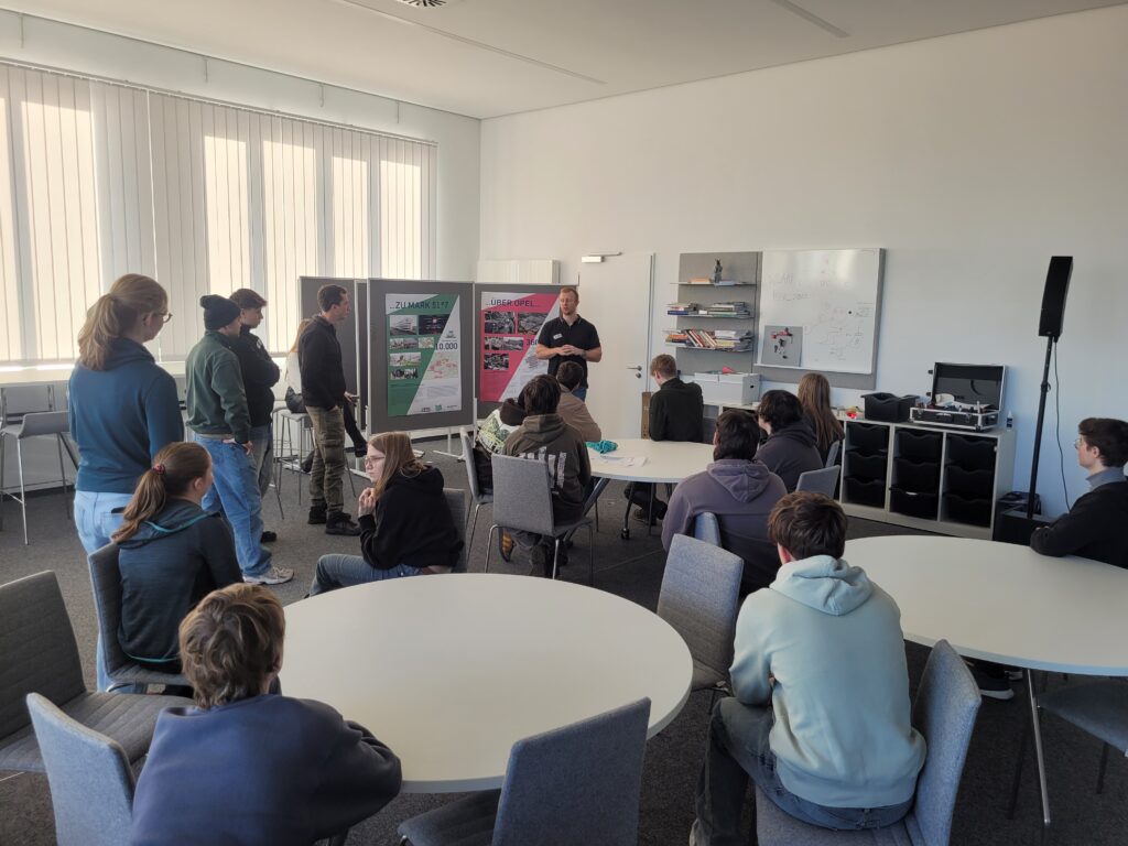 Eine Gruppe von Menschen sitzt und steht in einem hellen Klassenzimmer und hört einer Person zu, die Informationen auf großen Tafeln im vorderen Teil des Raums präsentiert.