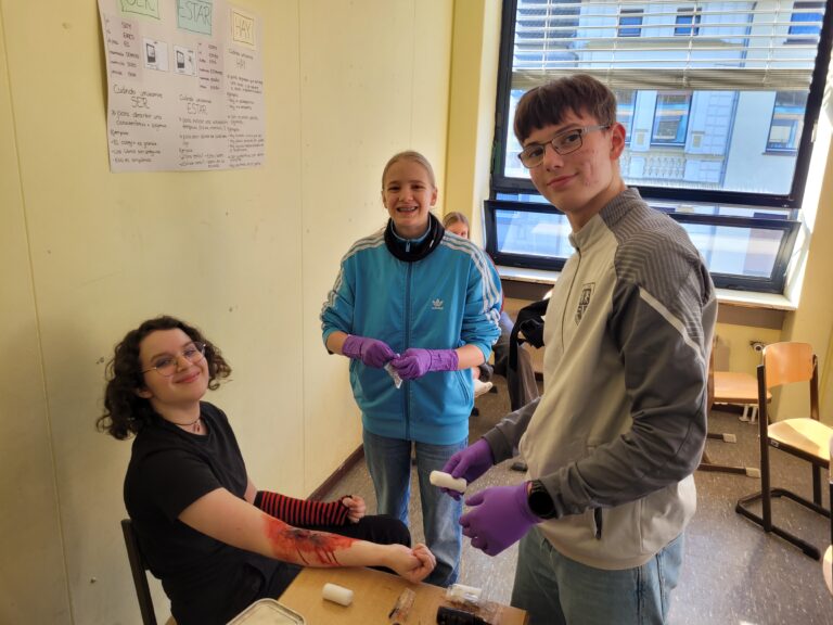 Drei Schüler üben in einem Klassenzimmer die Erste Hilfe an einer realistisch nachgebildeten Armverletzung; zwei stehen mit Handschuhen, einer sitzt lächelnd mit bandagiertem Arm da.