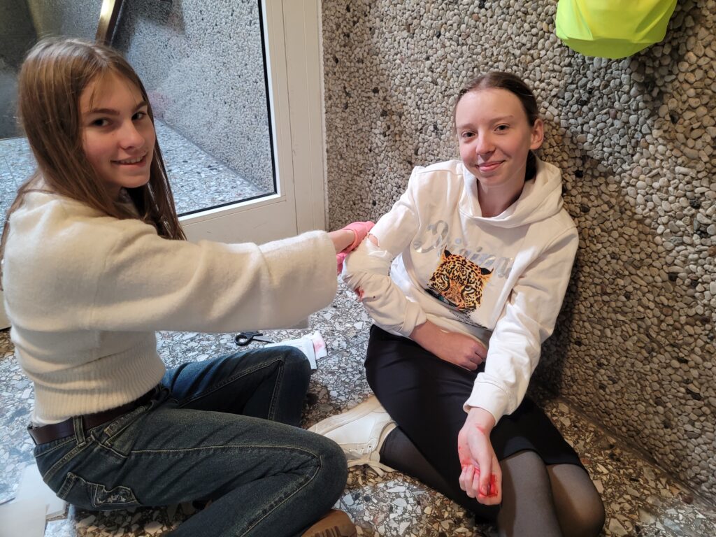 Zwei Mädchen sitzen auf dem Boden; die eine tut so, als würde sie der anderen Erste Hilfe am Arm leisten, der mit Kunstblut verschmiert ist. Beide lächeln in einem hellen Innenraum.