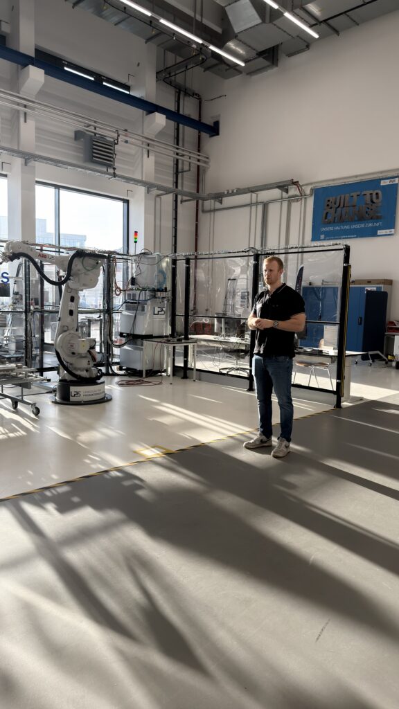 Ein Mann steht in einem hellen Industrielabor mit großen Fenstern, neben einem Roboterarm und verschiedenen Geräten, und das Sonnenlicht fällt auf den Boden.