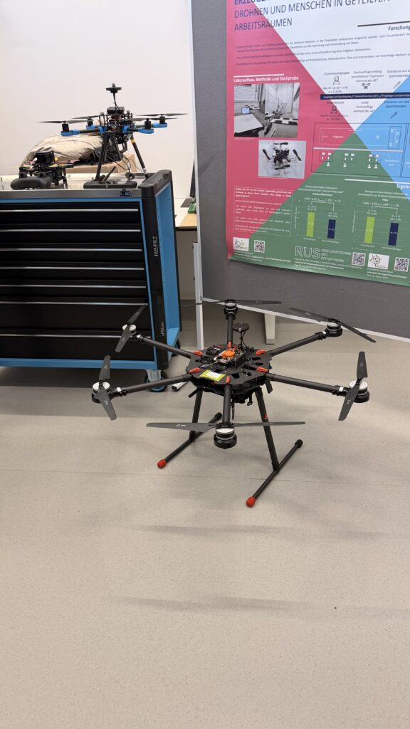 Eine große schwarze Drohne mit sechs Propellern liegt auf dem Boden neben einem Werkzeugschrank, während im Hintergrund eine weitere Drohne und ein technisches Poster zu sehen sind.