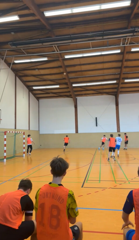Hallenfußballspiel in einer Turnhalle; Spieler in orangefarbenen und blauen Trikots auf dem Spielfeld, andere sitzen auf der Bank, darüber eine Holzdecke.