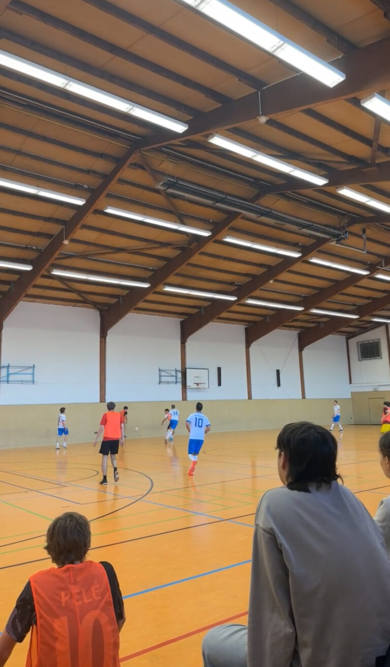 Eine Gruppe von Jungen spielt in einer Turnhalle Hallenfußball, während ein paar andere am Rand sitzen und das Spiel beobachten. Die Spieler tragen blau-weiße oder orangefarbene Trikots.