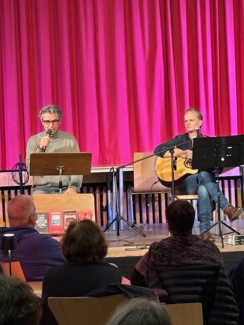 Zwei Männer treten auf der Bühne auf; einer spricht in ein Mikrofon, während der andere Gitarre spielt. Ein Publikum schaut zu. Im Hintergrund hängt ein rosa Vorhang.