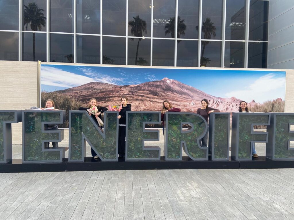 Sechs Personen stehen hinter großen "TENERIFE"-Schriftzügen vor einem Hintergrund, der eine Berglandschaft zeigt, vor einem modernen Gebäude mit Glasfenstern.