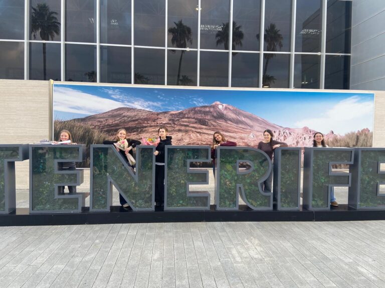 Sechs Personen stehen hinter großen "TENERIFE"-Schriftzügen vor einem Hintergrund, der eine Berglandschaft zeigt, vor einem modernen Gebäude mit Glasfenstern.