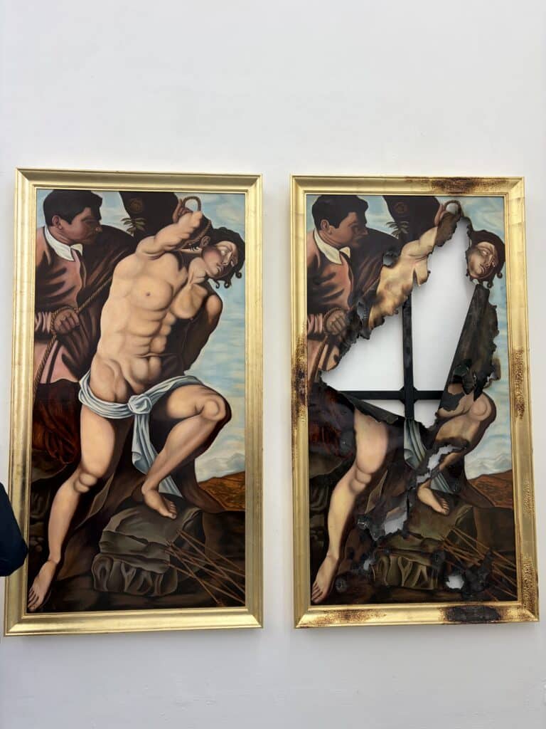 Zwei gerahmte Gemälde zeigen einen teilweise nackten Mann, der gefoltert wird; das rechte Gemälde ist beschädigt, wobei große verbrannte und zerrissene Teile die Wand dahinter freilegen.