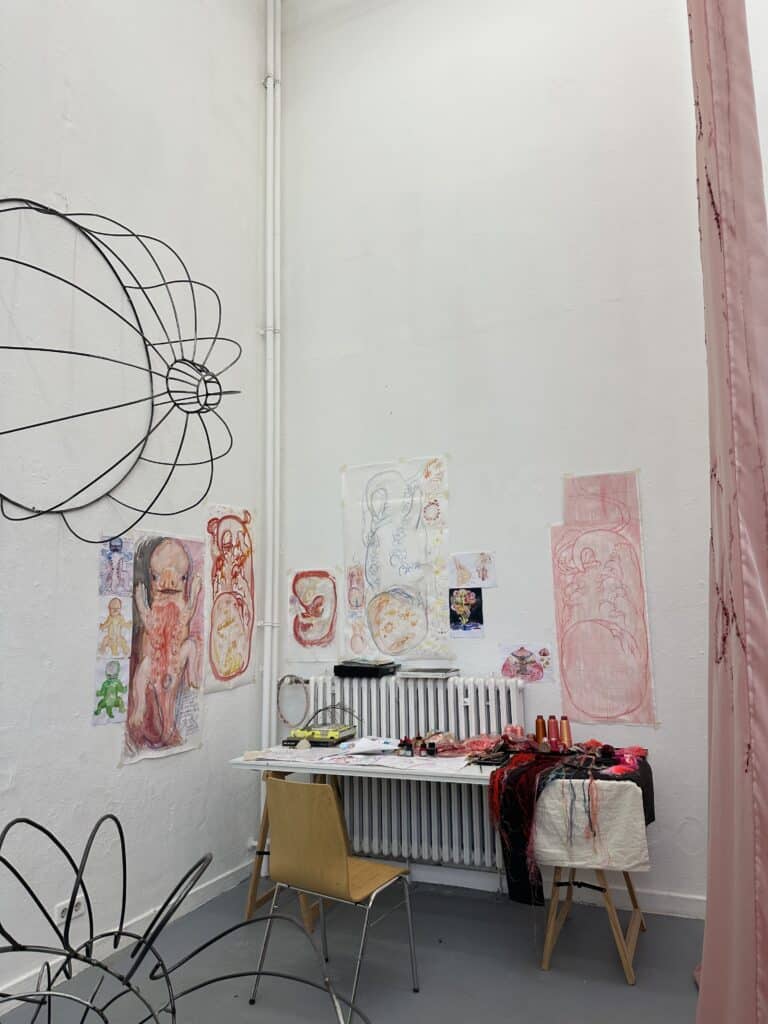 Eine Atelierecke mit einem Holzstuhl, einem Tisch mit Kunstzubehör und bunten, abstrakten Zeichnungen von menschlichen Figuren an weißen Wänden. Auch Drahtskulpturen sind zu sehen.