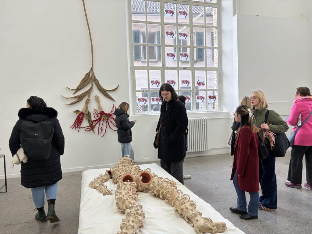 Die Besucher betrachten eine Kunstausstellung mit der Skulptur einer liegenden Figur mit hohlen Röhren am Körper, mit abstrakter Wandkunst und großen Fenstern im Hintergrund.