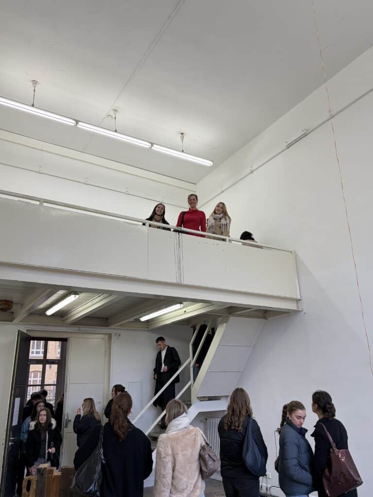 Menschen stehen und unterhalten sich in einem weißen, modernen Raum mit zwei Ebenen. Drei Frauen stehen auf der oberen Ebene, während andere unten in der Nähe einer Treppe stehen und sich unterhalten.