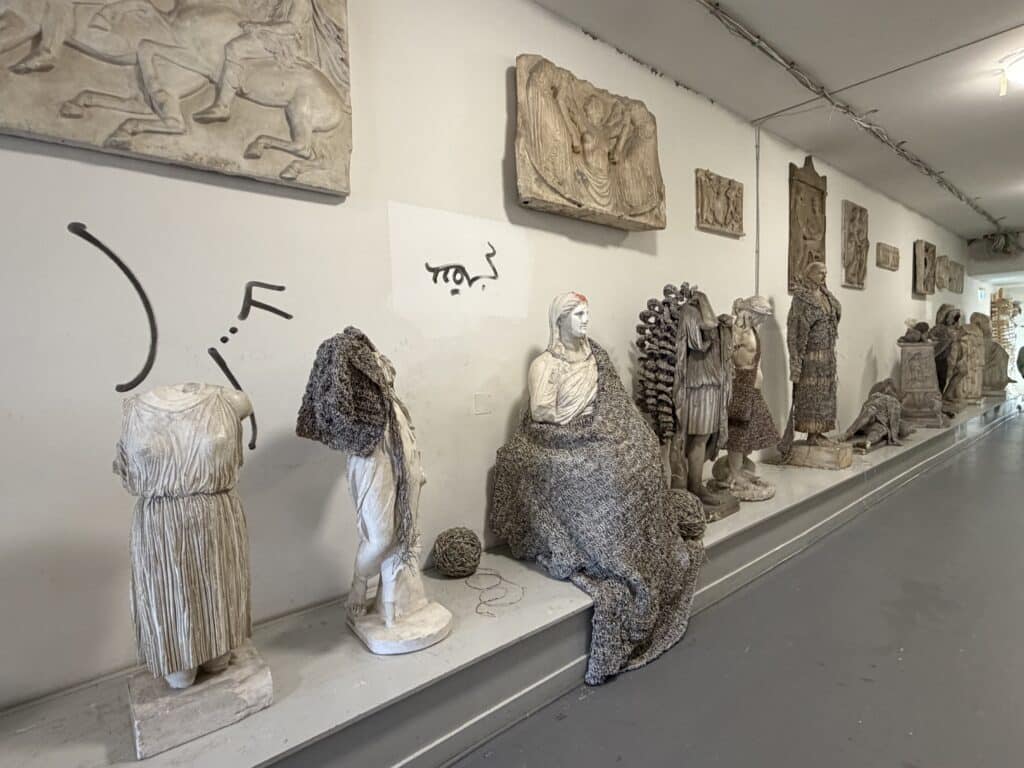 Eine Reihe von Skulpturen und Büsten im klassischen Stil, einige davon mit Netzen umhüllt, sind entlang einer Wand ausgestellt, darüber befinden sich Steinreliefs und abstrakte Markierungen.