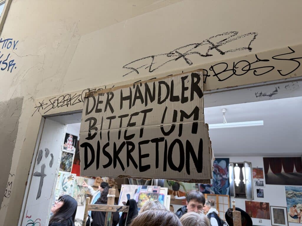 Ein handgeschriebenes Schild mit der Aufschrift "Der Händler bittet um Diskretion" hängt über einem mit Kunst gefüllten Raum.