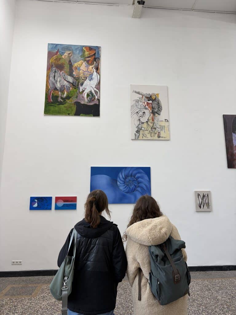 Zwei Personen mit Rucksäcken stehen vor einer weißen Wand, an der mehrere bunte und abstrakte Gemälde in einer Kunstgalerie hängen.