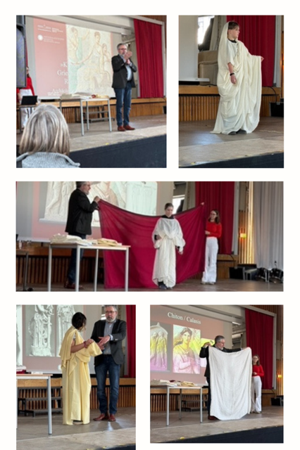 Ein Redner auf der Bühne stellt sich dem Publikum vor, während Menschen in einem Klassenzimmer oder einer Vorlesung Kleidungsstücke im antiken griechischen Stil mit Stoff modellieren.