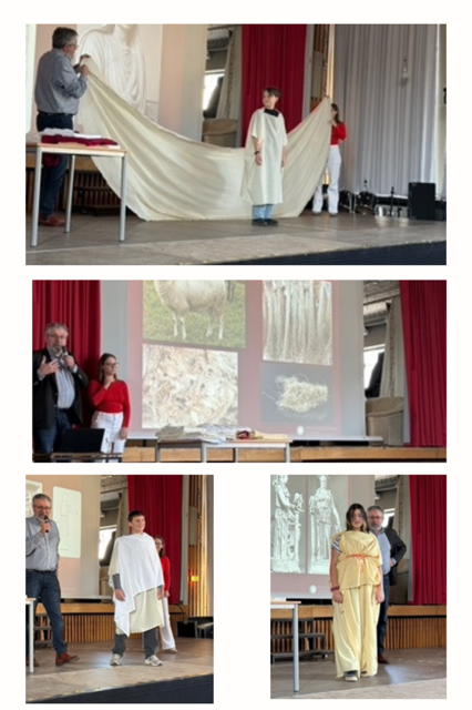 Ein Mann drapiert weißen Stoff an einer Schülerin, während andere assistieren; später modellieren zwei Schüler auf der Bühne Kleidungsstücke im antiken Stil mit Bildern von Wolle und Stoff hinter ihnen.