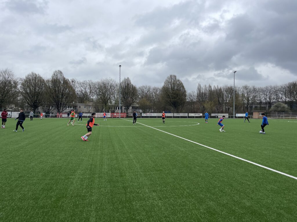 Mehrere Personen spielen Fußball auf einem grünen Feld unter einem bewölkten Himmel, mit Bäumen und Gebäuden im Hintergrund.