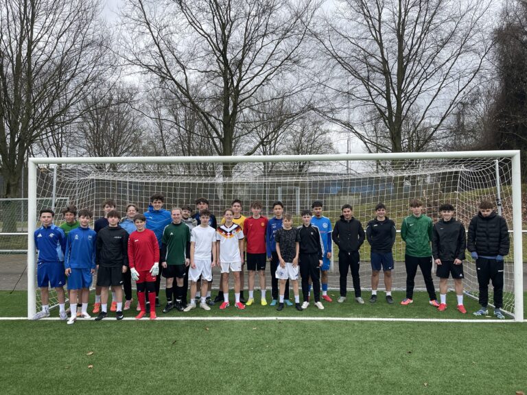Eine Gruppe von Jungen in Sportkleidung steht zusammen vor einem Fußballtor auf einem Feld und posiert für ein Mannschaftsfoto an einem bewölkten Tag mit Bäumen im Hintergrund.