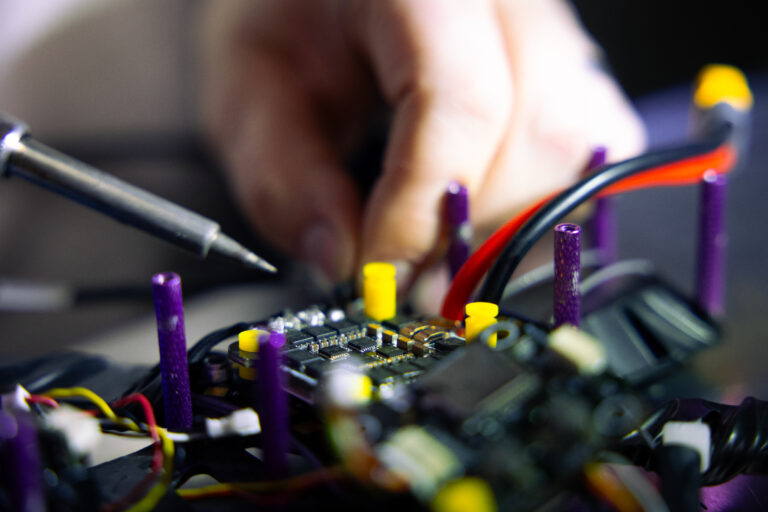 Nahaufnahme einer Hand beim Löten elektronischer Bauteile auf einer Leiterplatte, wobei bunte Drähte und Stifte sichtbar sind.
