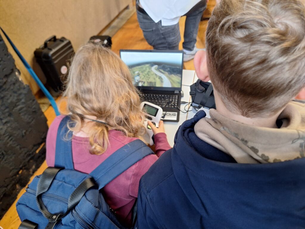 Zwei Kinder schauen auf einen Laptop, auf dem eine Drohne aus der Luft zu sehen ist. Ein Kind hält einen Controller, während in der Nähe Ausrüstungskoffer auf dem Boden zu sehen sind.