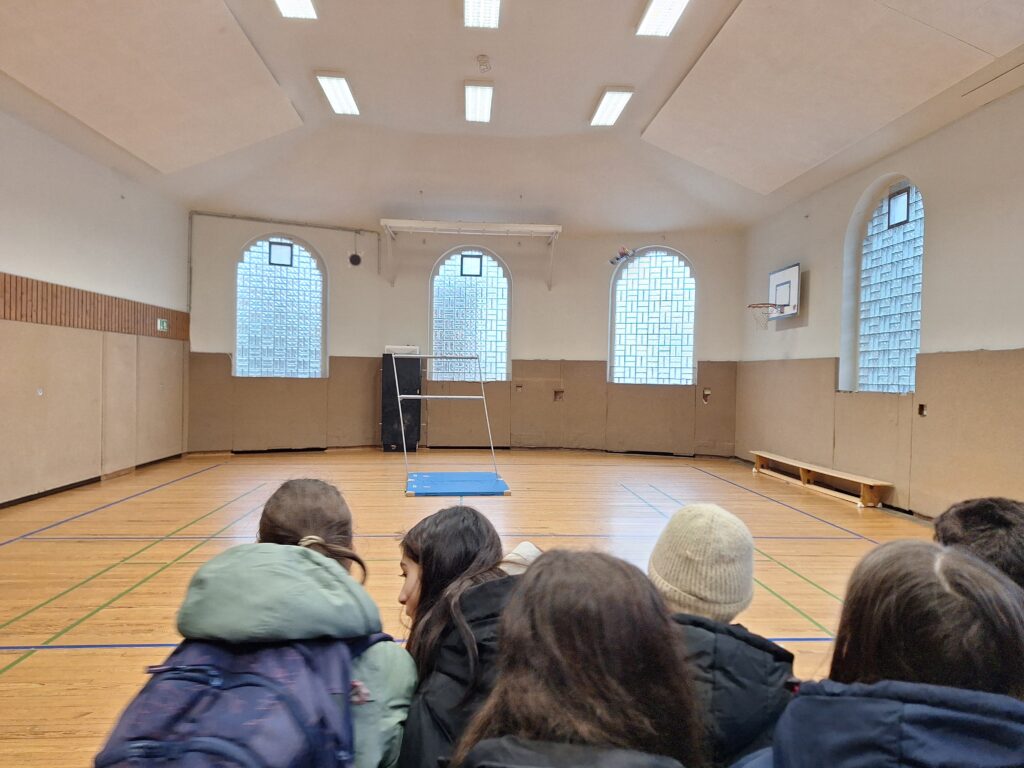 Eine Gruppe von Schülern sitzt in einer Turnhalle auf dem Boden, vor einer blauen Matte und Sportgeräten in der Nähe großer Fenster mit Glasbausteinen.
