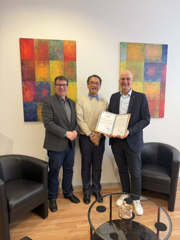 Drei Männer stehen lächelnd in einem Raum, einer hält eine offene Urkunde in der Hand. Hinter ihnen hängen zwei abstrakte bunte Gemälde an der Wand. Zwei schwarze Stühle stehen in der Nähe.