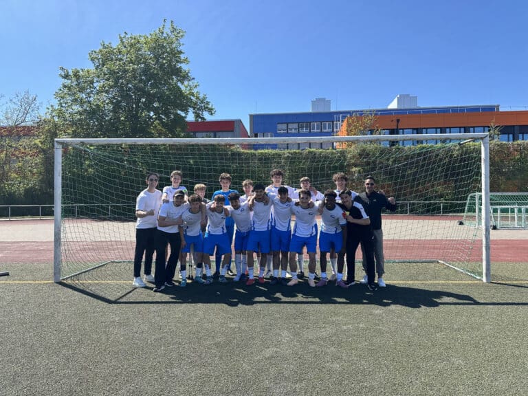 Eine Fußballmannschaft von Jungen im Teenageralter posiert gemeinsam vor einem Tor auf einem Feld im Freien mit klarem Himmel und bunten Gebäuden im Hintergrund.