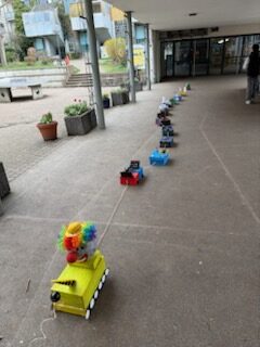 Eine Reihe bunter, selbstgebauter Spielzeugfahrzeuge, angeführt von einem gelben Fahrzeug mit einer Clownsfigur, steht draußen in der Nähe eines Gebäudeeingangs.