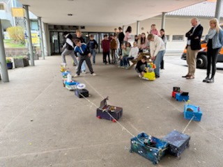 Kinder und Erwachsene sehen zu, wie ein Junge kreative, bunte, selbstgebaute Karren zieht, die mit einer Schnur vor einem Gebäude unter einem überdachten Gehweg verbunden sind.