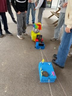 Eine Gruppe von Menschen steht um drei kleine, bunte, selbst gebaute Autos herum, die mit Schnüren auf einem Betonboden verbunden sind.