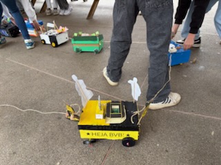 Kleine handgefertigte Autos, darunter ein gelbes mit der Aufschrift HEJA BVB, werden von Menschen auf dem Boden arrangiert, von denen einige in der Nähe hocken oder stehen.