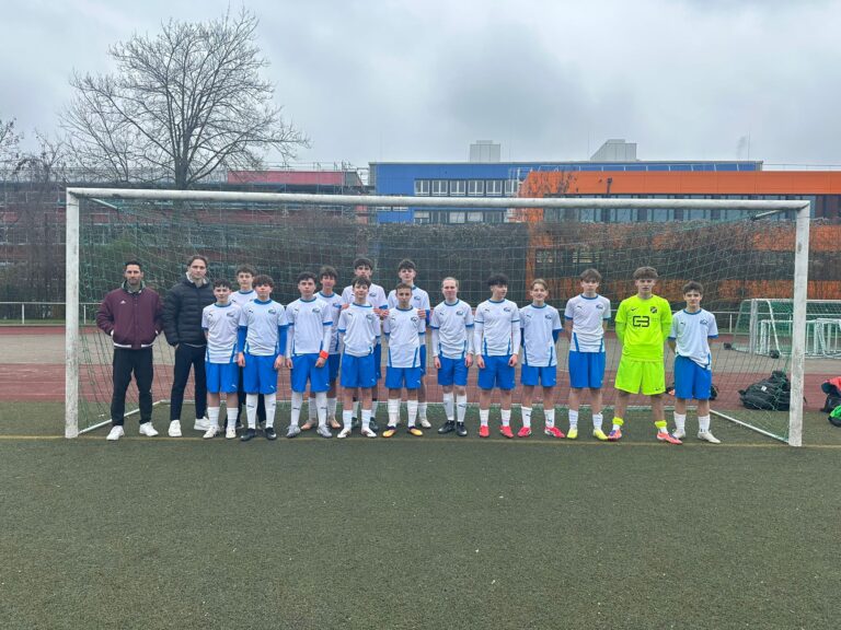 Eine Jugendfußballmannschaft in weiß-blauen Trikots steht an einem bewölkten Tag zusammen mit zwei Trainern vor einem Tor, im Hintergrund sind Schulgebäude zu sehen.