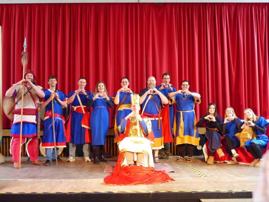 Eine Gruppe von Menschen in farbenfrohen mittelalterlichen Kostümen steht auf einer Bühne mit roten Vorhängen und bildet mit ihren Händen Herzformen; eine Person sitzt in der Mitte auf einem Thron.