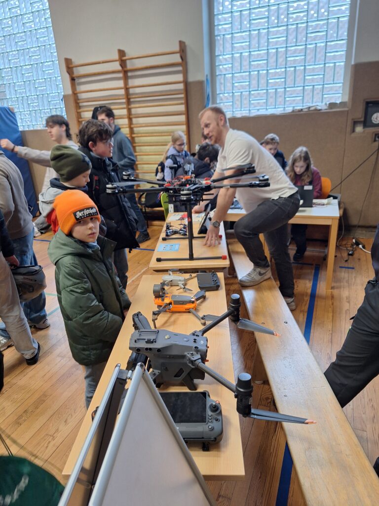In einer Sporthalle versammeln sich Menschen um Tische, auf denen verschiedene Drohnen und Steuergeräte ausgestellt sind. Ein Mann erklärt Kindern und Erwachsenen die Geräte.