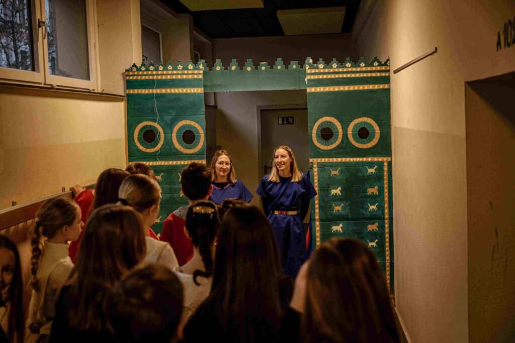 Zwei Frauen in blauen Kleidern stehen vor einer Gruppe von Kindern unter einem dekorativen Torbogen mit großen Augen und Tiermotiven in einem Flur.