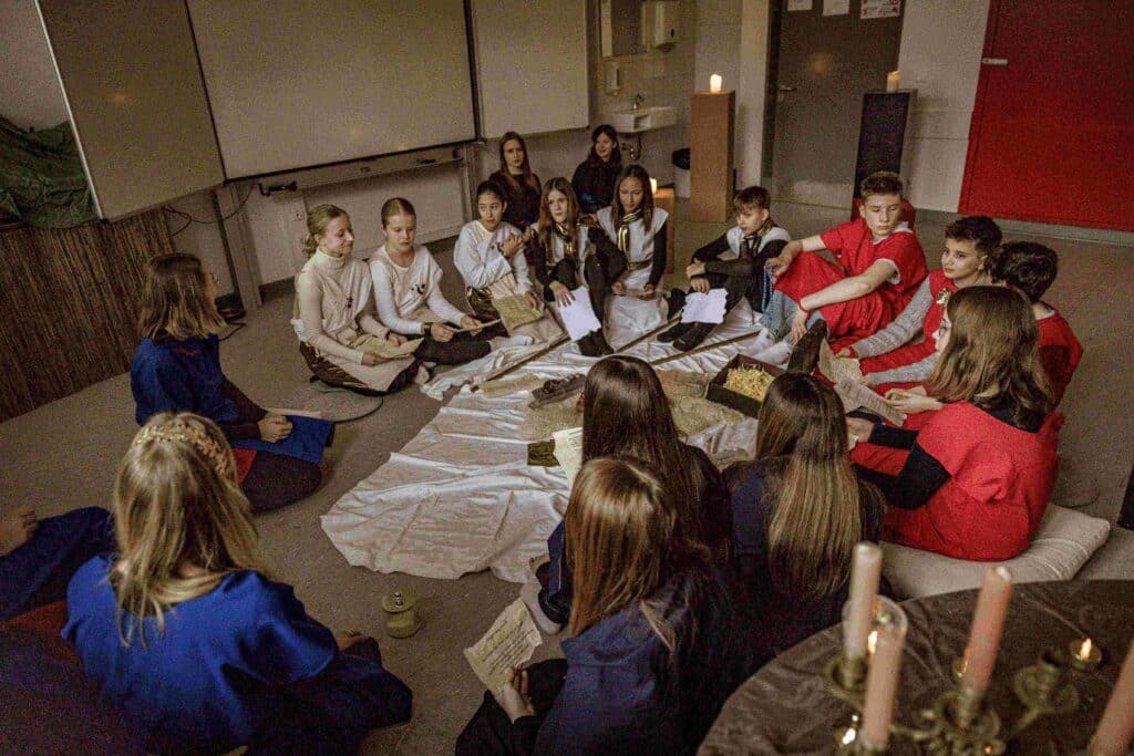 Eine Gruppe kostümierter Kinder sitzt in einem Kreis auf dem Boden, hält Papiere in der Hand und sitzt mit Kerzen und Requisiten in einem schwach beleuchteten Klassenzimmer.
