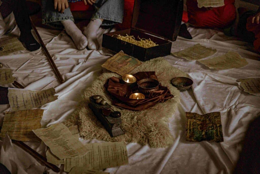 Eine Gruppe von Menschen sitzt im Kreis auf einem weißen Tuch, umgeben von Kerzen, alten Papieren, einer Kiste mit Stroh und alten Gegenständen, die eine geheimnisvolle Atmosphäre schaffen.