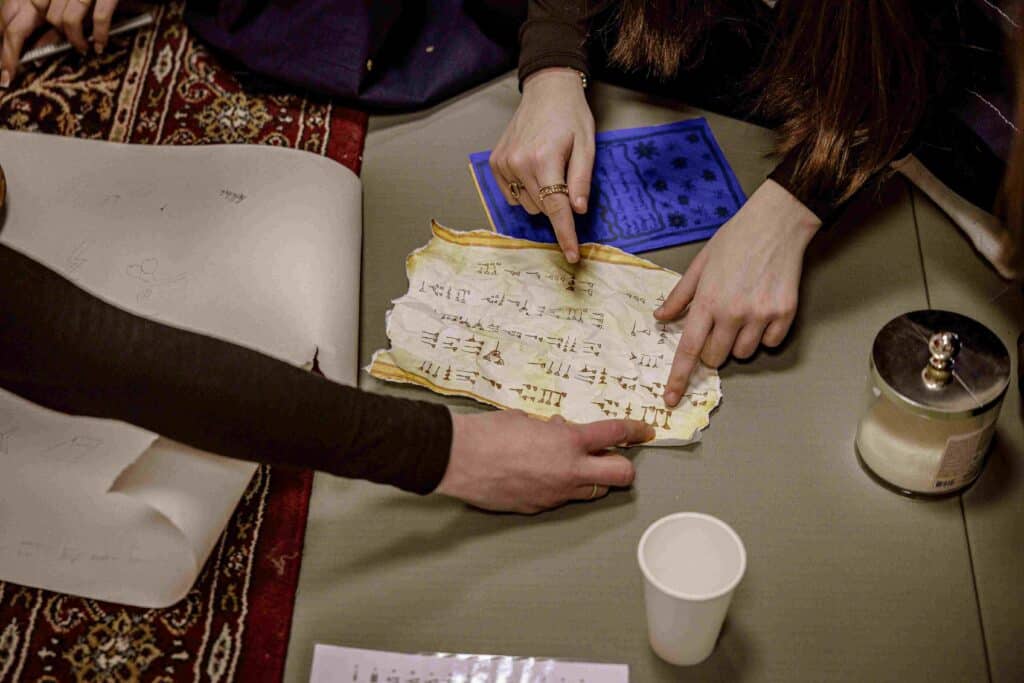 Drei Personen untersuchen ein altes, vergilbtes Pergament mit handschriftlichen Notizen, zeigen auf Symbole und Text, umgeben von Papieren, einer Kerze und einem Pappbecher.