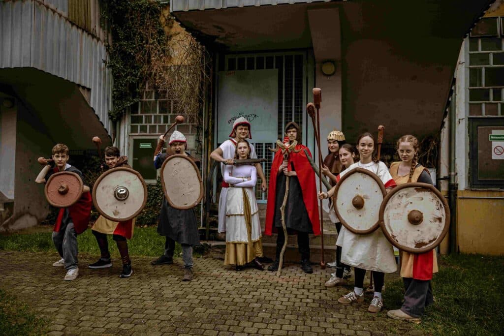 Eine Gruppe von Menschen in mittelalterlichen Kostümen posiert im Freien mit Schilden und Stäben vor einem Gebäude mit überwucherten Ranken und vergitterten Türen.
