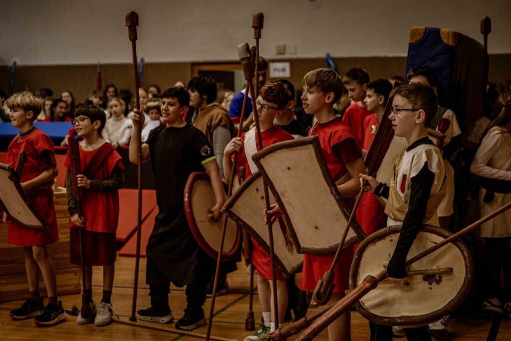 Eine Gruppe von Kindern in Kostümen mit Schilden und Stäben, die sich in einem Raum versammelt haben, als ob sie sich auf eine Scheinschlacht oder eine historische Nachstellung vorbereiten.