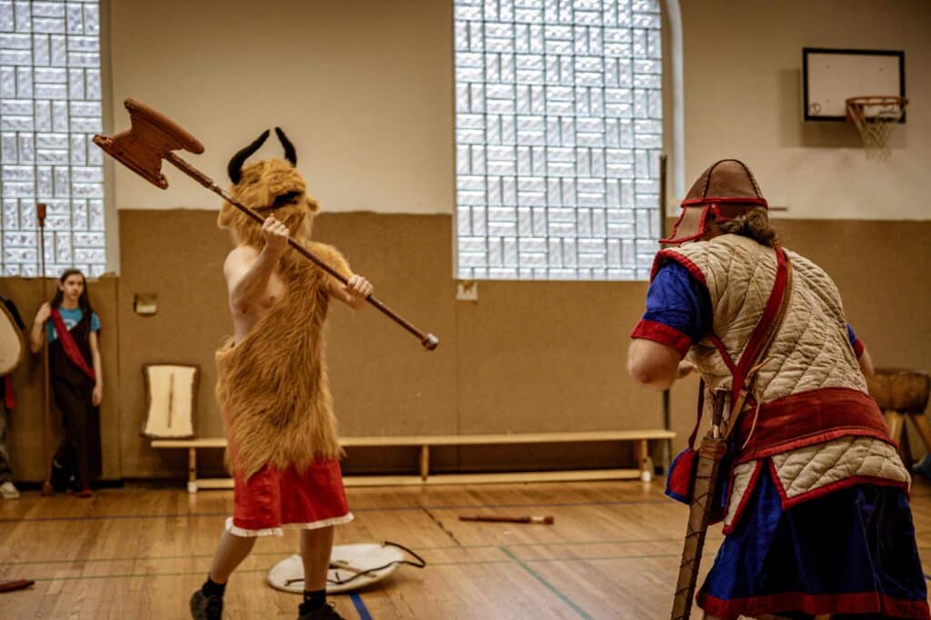 Zwei Personen in historischen Kostümen bereiten sich in einer Turnhalle auf ein Duell mit hölzernen Waffen vor; die eine trägt einen gehörnten Pelzanzug, die andere ein rot-blau gepolstertes Kostüm.