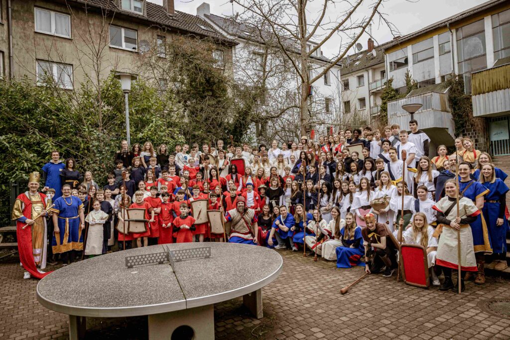 Eine große Gruppe von Menschen in mittelalterlichen Kostümen posiert gemeinsam im Freien in der Nähe einer Tischtennisplatte in einem Wohnhof.