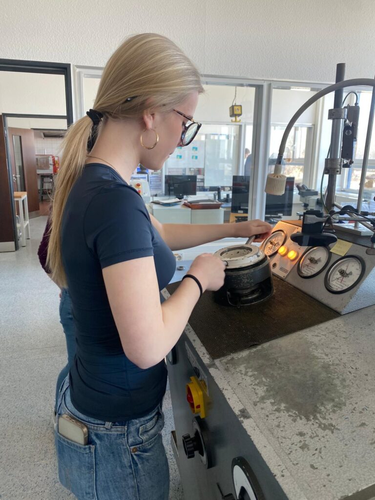 Eine Frau mit langen blonden Haaren und einer Brille bedient in einem hellen, modernen Labor eine Maschine mit Zifferblättern und Messinstrumenten.